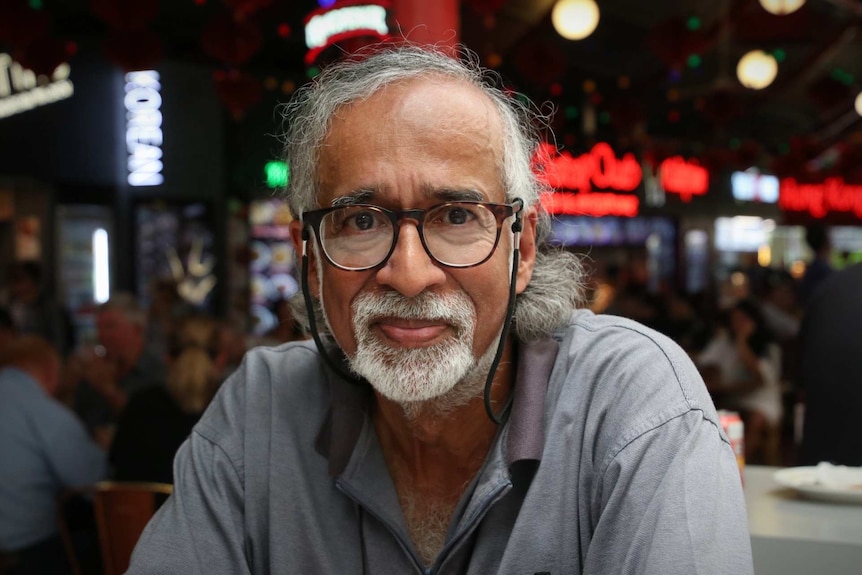 A man with a greying beard and glasses sits in a busy food hall