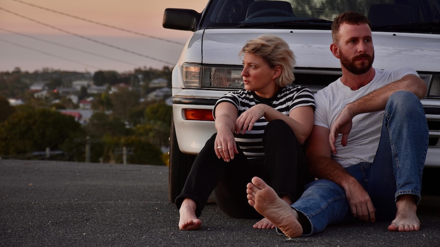 Tracy McNeil and Dan Parsons sit on the road in front of a white sedan. They are barefoot and leaning on each other.
