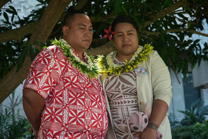 Two people wearing leis stand under a tree, looking sad.