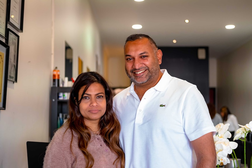 You view two people of South Asian descent looking into the camera as they stand in front of a shelf carrying hair products.