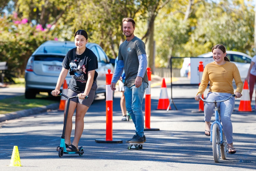 A teenage girl on a scooter, a man on a skateboard and teenage girl riding a bike down a leafy Woodridge street