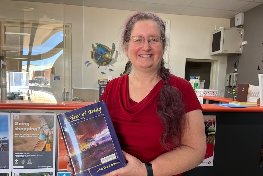 A woman with long hair, wearing a red top, in a library holding a book.