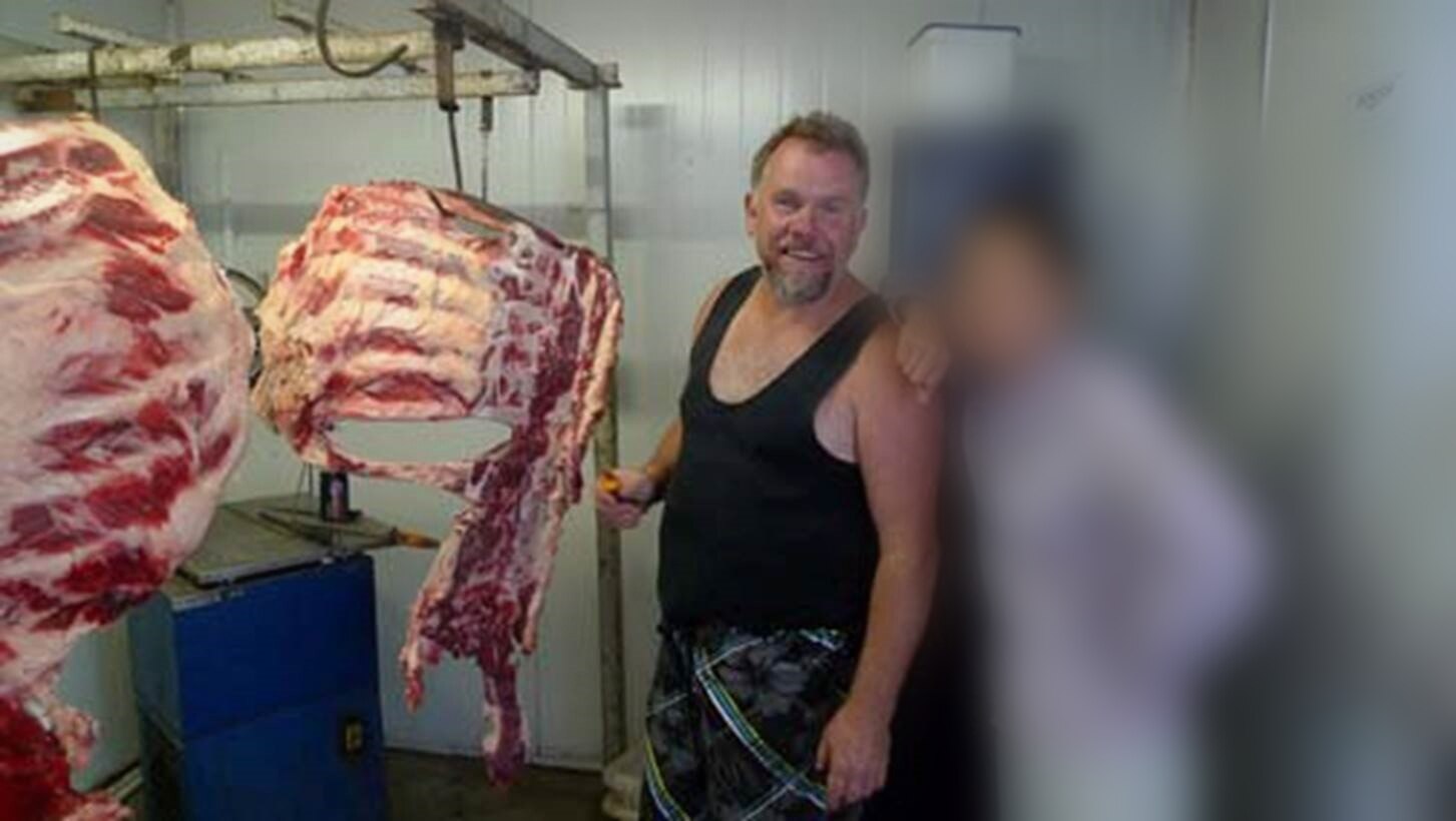 A man and person with a blurred face next to hanging horse carcasses.