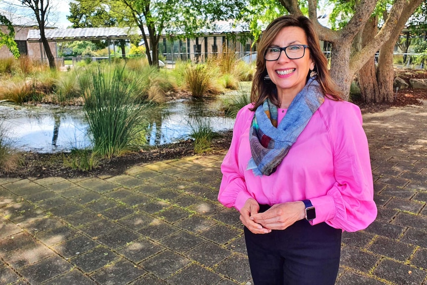 A woman in a pink blouse and blue scarf smiles in front of a pond area.