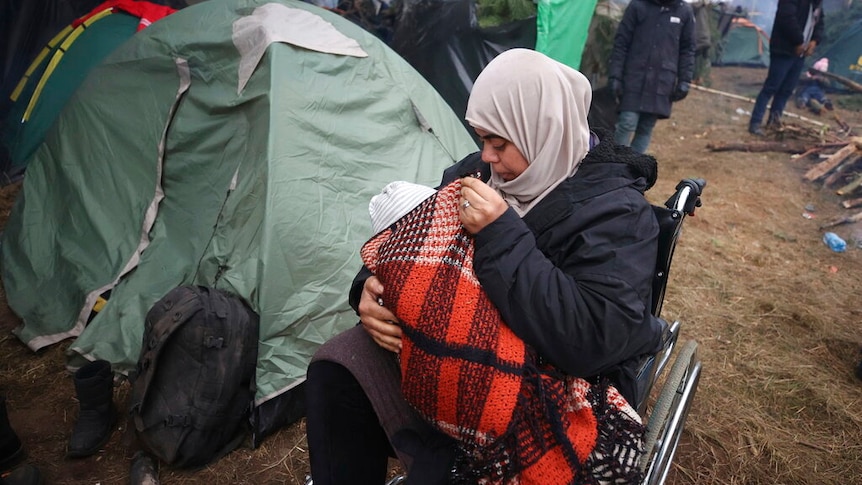 A woman holds her child as she sits in a wheelchair as other migrants gather at Belarus-Poland border.