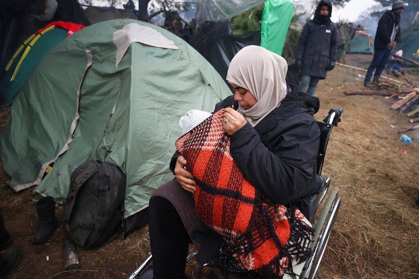 A woman holds her child as she sits in a wheelchair as other migrants gather at Belarus-Poland border.