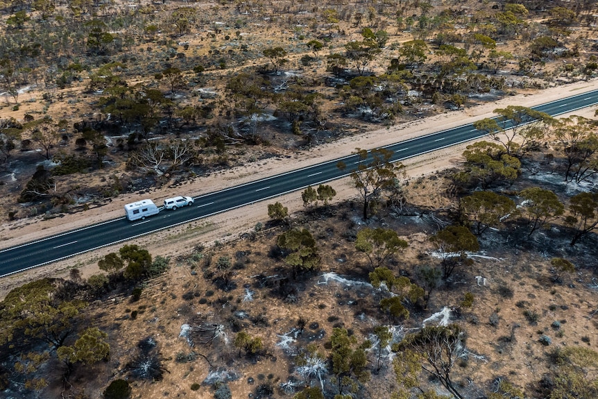 A drone photo above the highway with bush burnt on either side and a car and caravan travelling through