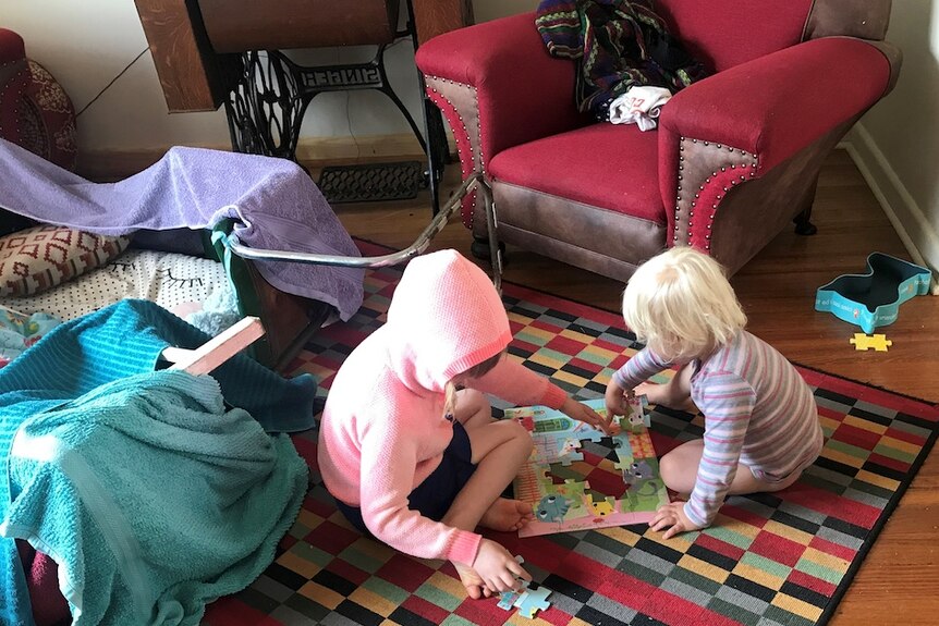 Two young children sit on the floor of a lounge room and play with toys.