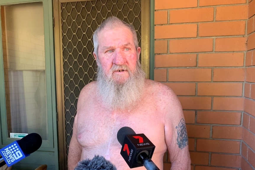 An old man with a beard and no shirt speaking in front of microphones