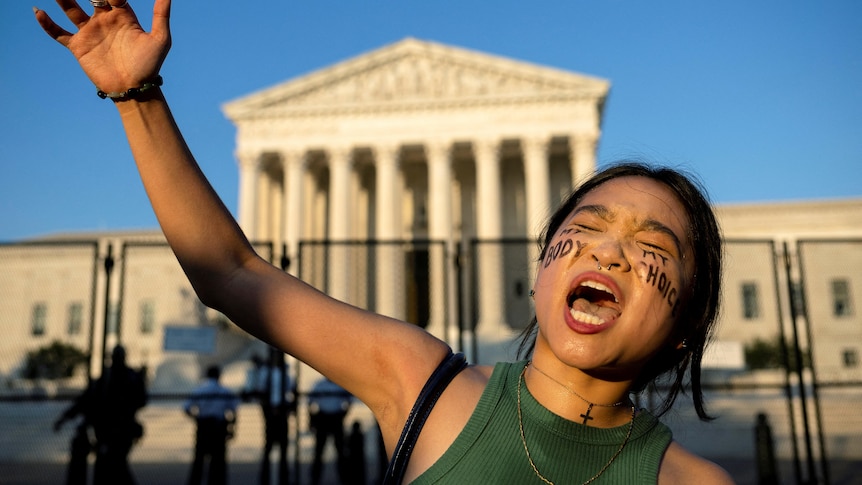 A young woman outstretches her arm as she yells outside the Supreme Court. She has 'my body, my choice' written on her cheeks