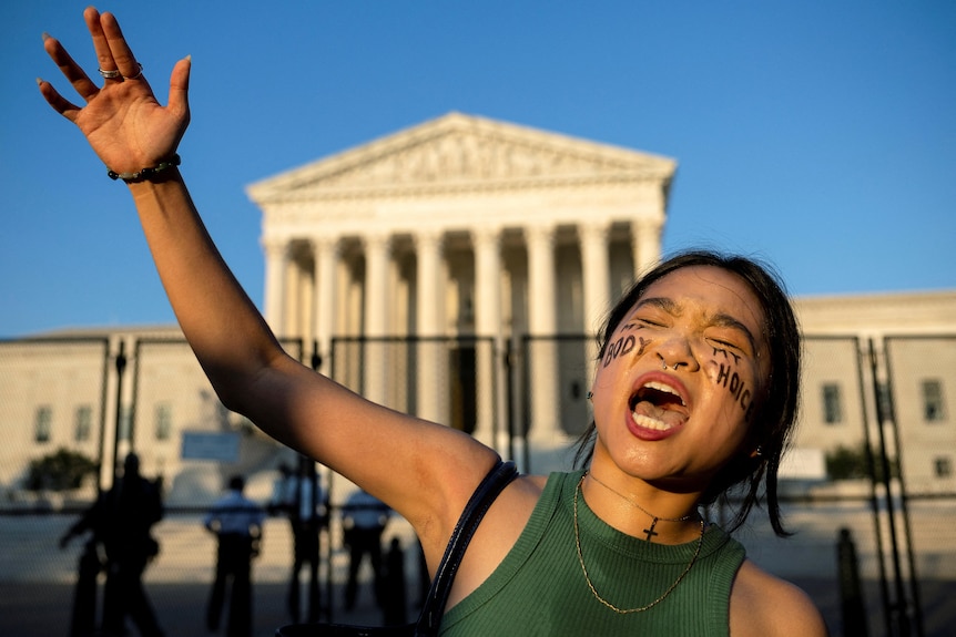 A young woman outstretches her arm as she yells outside the Supreme Court. She has 'my body, my choice' written on her cheeks