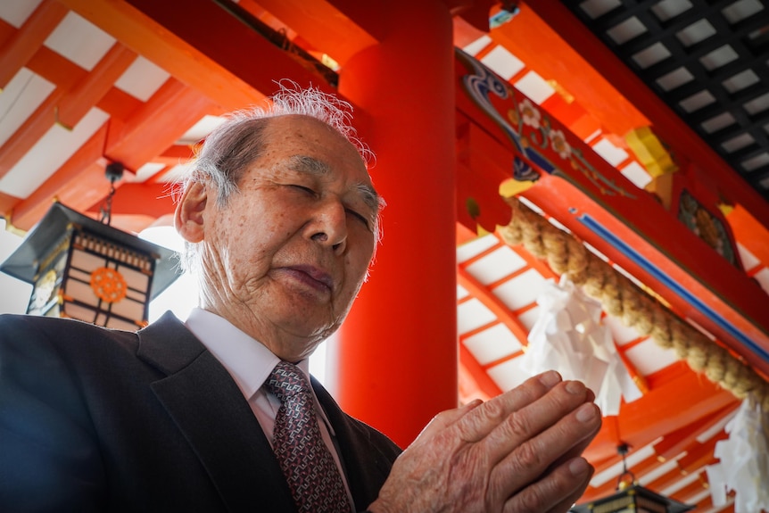 An older Japanese man with his eyes closed and palms pressed in prayer