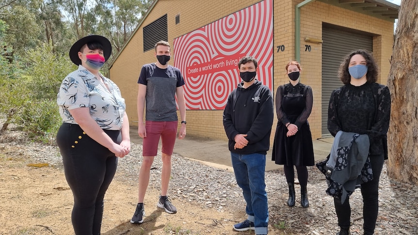 Five masked people stand in front of a building with a poster reading "Create a world worth living"