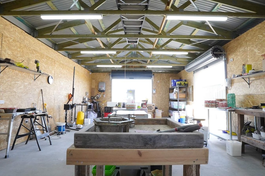 Work benches, shelving and tools fill a large work shed with lots of natural sunlight entering through open roller doors.