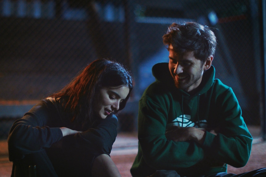 A 20-something woman and man in hoodies sitting close to each other outside at night, smiling.