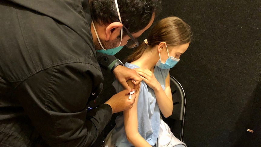 A male nurse gives a vaccine to a girl wearing a face mask.