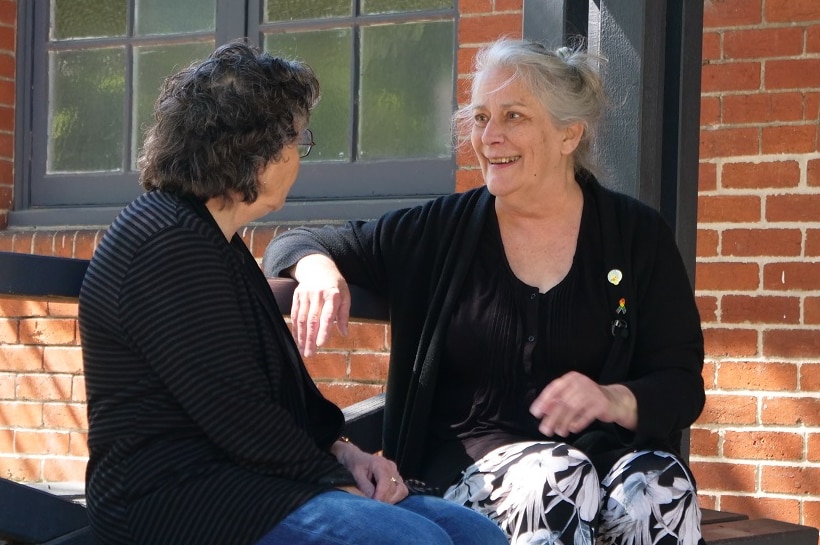 Maurya Bourandanis is dressed in black as she smiles and speaks to someone sitting next to her on a bench.