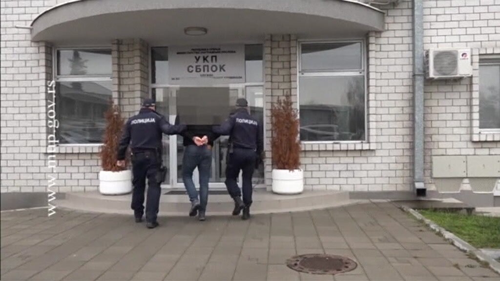 Two police lead man wearing jeans and a black jacket with his arms handcuffed behind his back into a government building