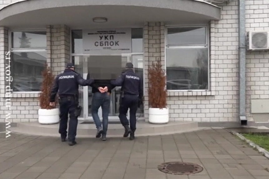 Two police lead man wearing jeans and a black jacket with his arms handcuffed behind his back into a government building