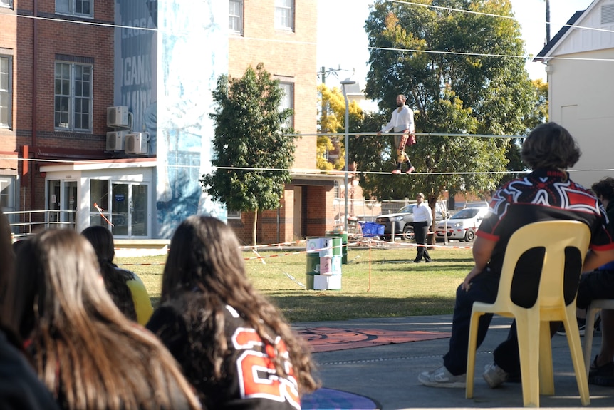 Students sitting on the ground watching a man walk on a wire