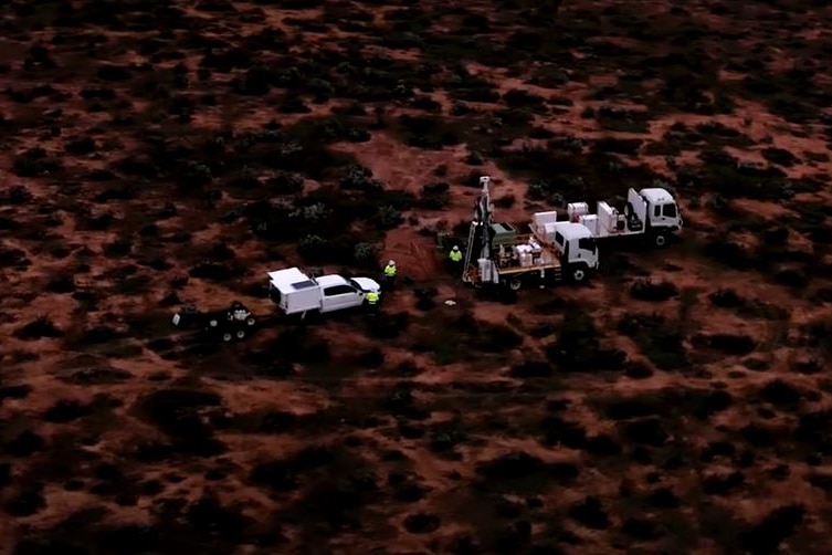 A drill rig surrounded by white vehicles in an arid environment, pictured from the air.