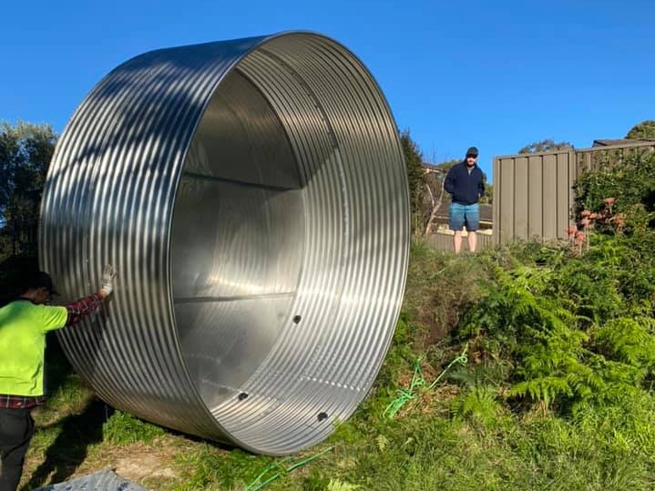 Man in Fluro rolls huge steel tank through hole in the fence. 