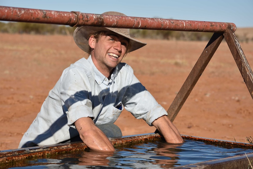 A man smiles wide with his arms elbow-deep in murky water at a cattle trough in an orange dirt paddock