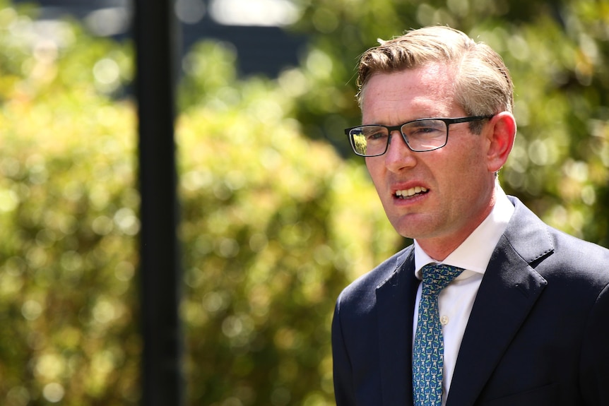 A man with strained expression on his face stands wearing a suit and glasses in front of a garden. In front of him is a woman