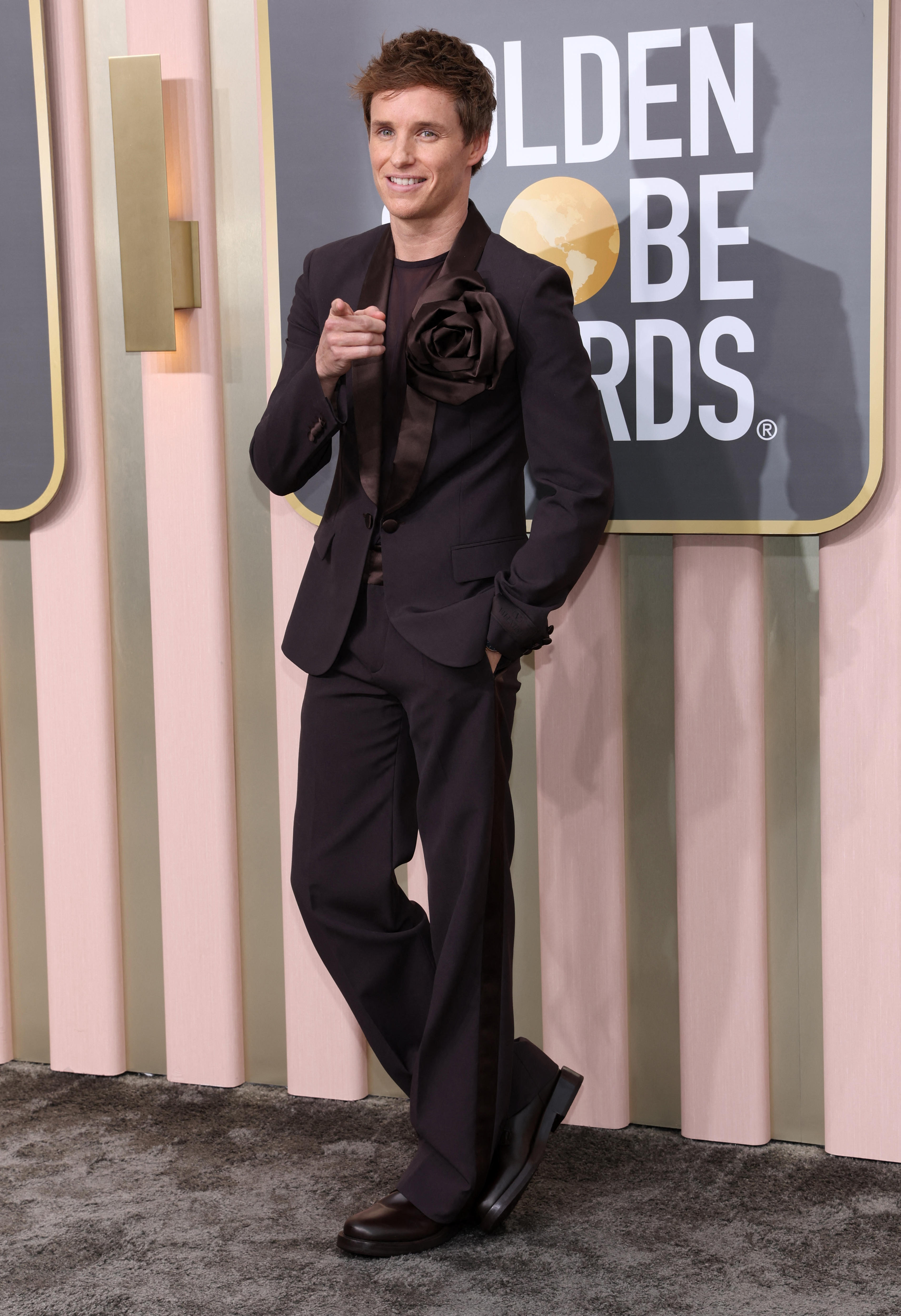 Eddie Redmayne wearing a dark brown silky suit with a rose detail on the jacket.