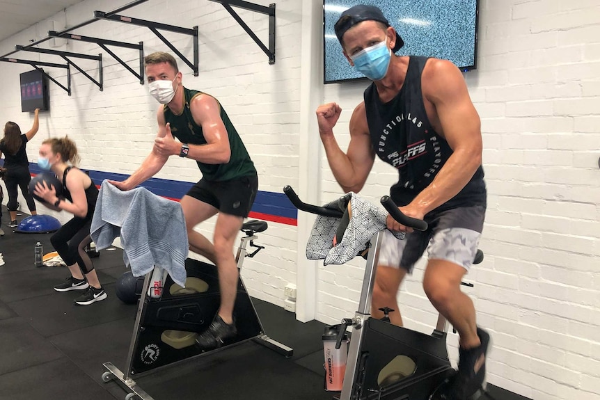 Two men in singlets wearing face masks give the thumbs up while riding exercise bikes at F45 training.