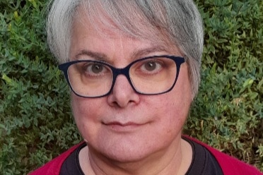 A woman with short hair wearing glasses, standing in front of a hedge.