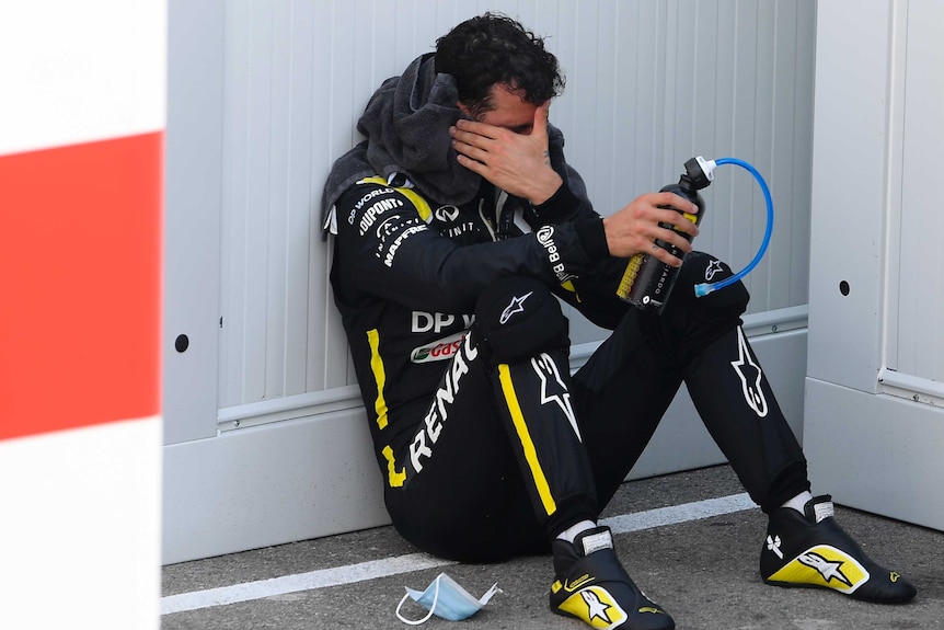 Daniel Ricciardo holds his head with one hand and a bottle in the other, sitting slumped next to a wall