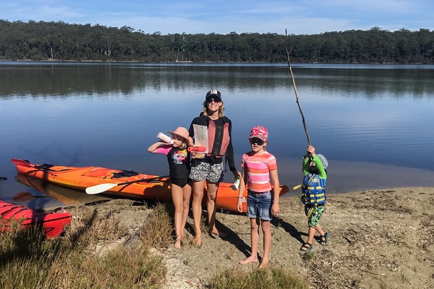 A woman with three small young children wearing hats stand by a pair of orange kayaks on a calm water's edge.