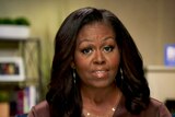 Michelle Obama in a brown top and gold hoop earrings speaking in a living room