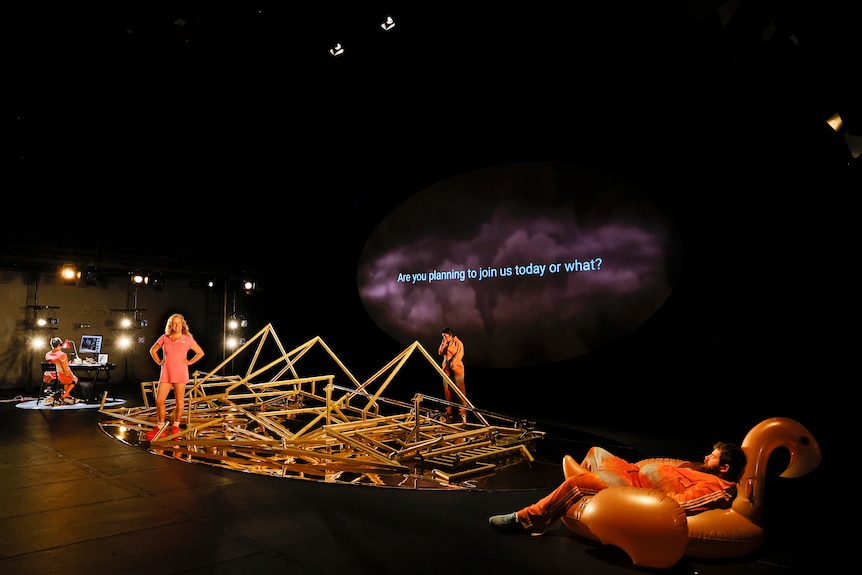 A man lounges in an inflatable flamingo while two women work on an abstract golden structure on a stage
