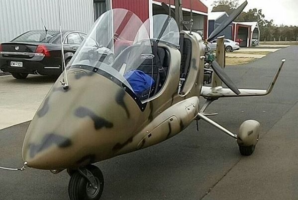 A gyrocopter sits on the tarmac with camouflage paintwork.