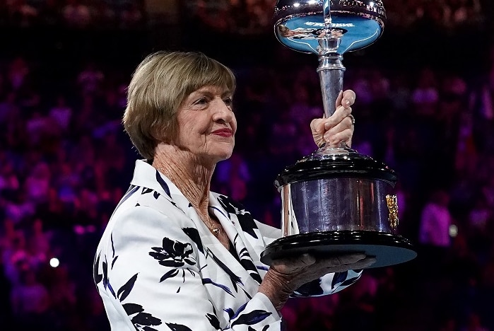 A woman holds a large trophy aloft under lights on a tennis court with a large number of spectators in the stands.