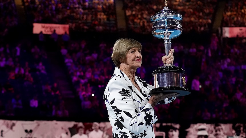 A woman holds a large trophy aloft under lights on a tennis court with a large number of spectators in the stands.