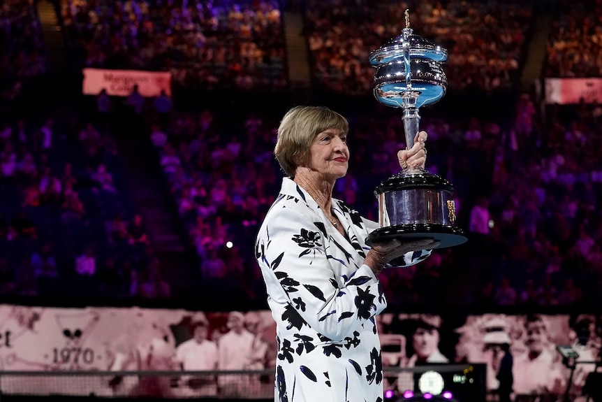 A woman holds a large trophy aloft under lights on a tennis court with a large number of spectators in the stands.