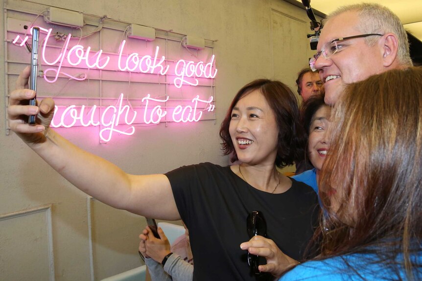 A woman holds a phone up in front of her, Scott Morrison and others. A sign on the wall says: "You look good enough to eat"