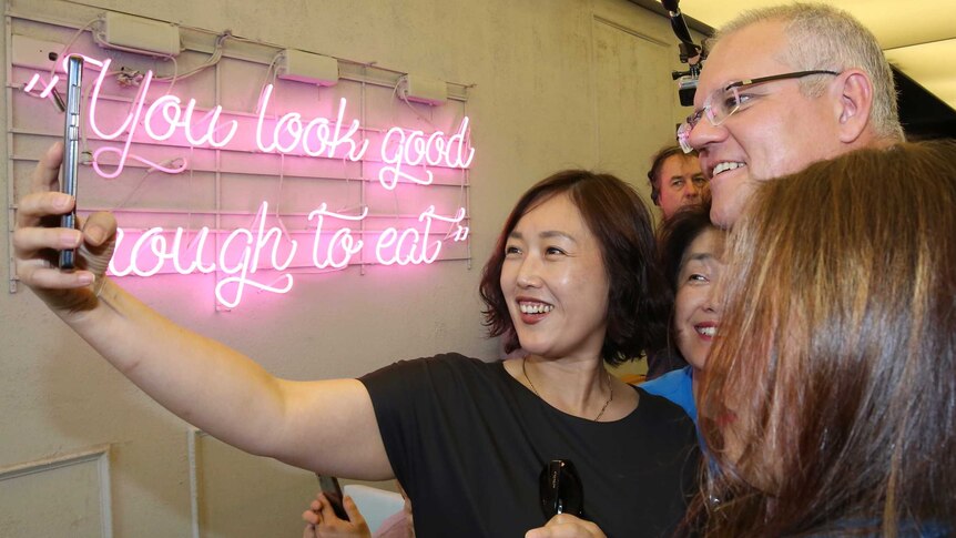 A woman holds a phone up in front of her, Scott Morrison and others. A sign on the wall says: "You look good enough to eat"