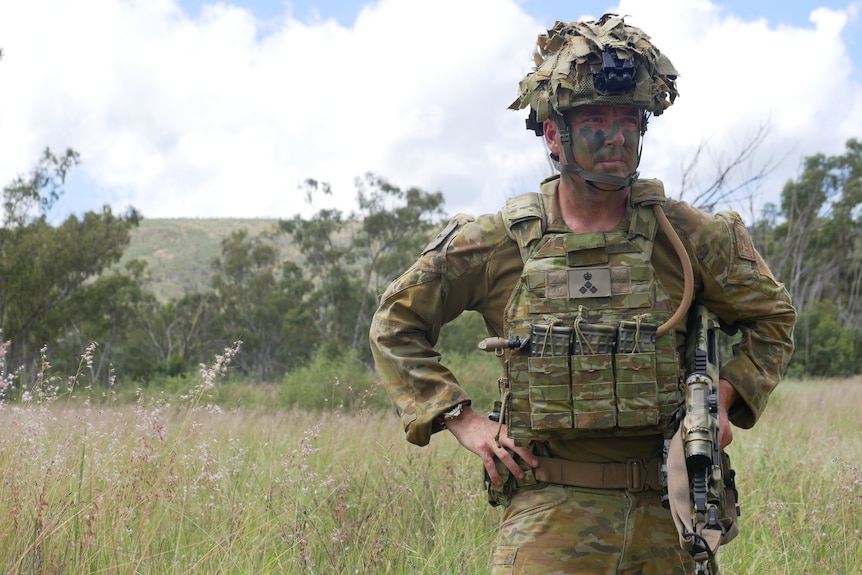 A man in camouflage stands in a field near bush scrub and trees