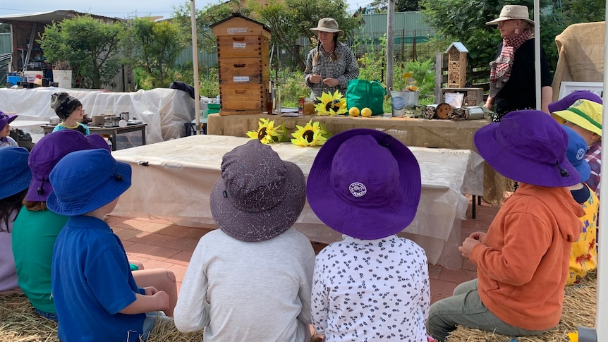 children sitting in hats with backs to the camera, in a circle outdoors, watching an instructor