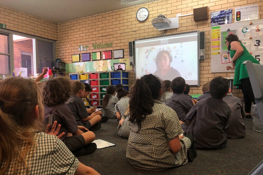 Students from St Francis Xavier  sit on the mat looking at a screen where teacher Regina Wan teaches them from Beijing.