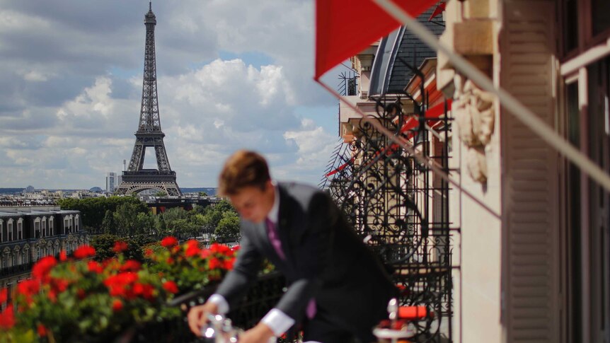 A man wearing a suit on a balcony in the foreground out of focus while the Eiffel Tower is in the background.