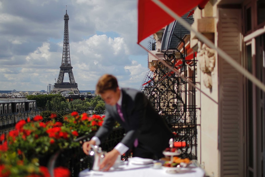 A man wearing a suit on a balcony in the foreground out of focus while the Eiffel Tower is in the background.