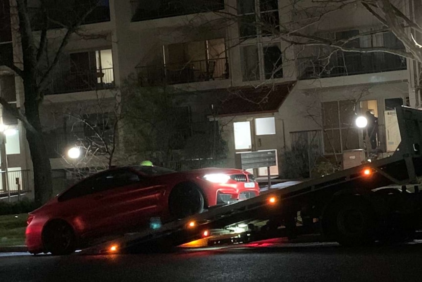 A red sedan is lifted onto a tow truck at night outside an apartment block