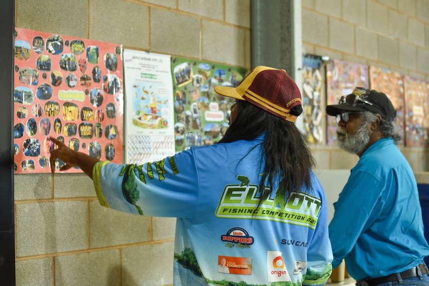 A woman points at a diagram on a posters on a brick interior wall. A man is listening.