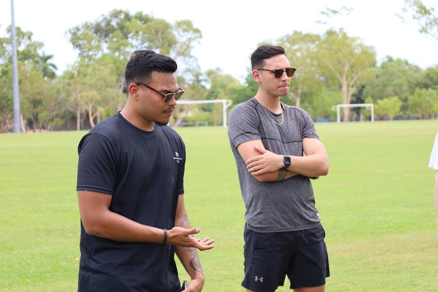 Two men are pictured, both are wearing sunnies are wearing dark shirts. They are standing on a field with soccer goals.