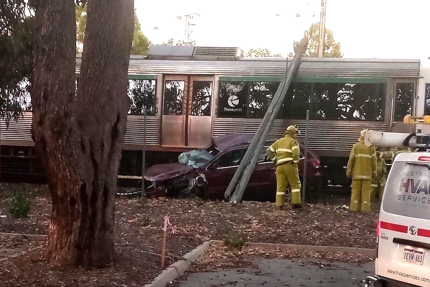 A side-on shot of the wreckage of a dark red car crushed net to a train with two firefighters nearby in yellow clothing.